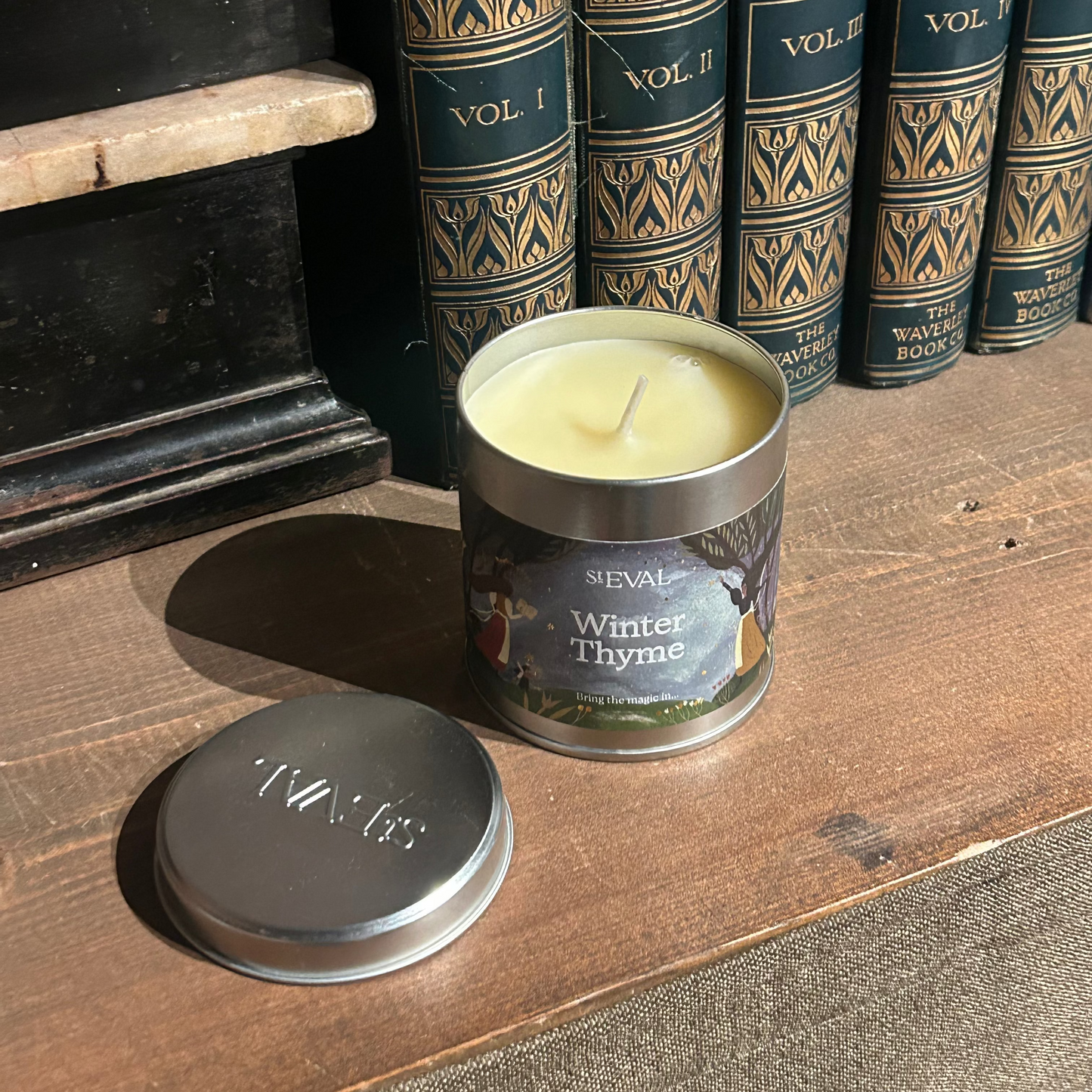 Candle in a metal tin labeled 'Winter Thyme' on a wooden surface with books in the background.