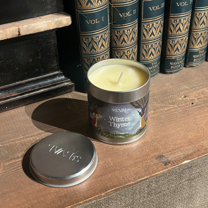 Candle in a metal tin labeled 'Winter Thyme' on a wooden surface with books in the background.